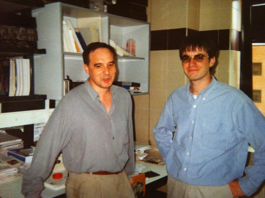 Dos hombres en un laboratorio, conversando, rodeados de libros y equipo científico.