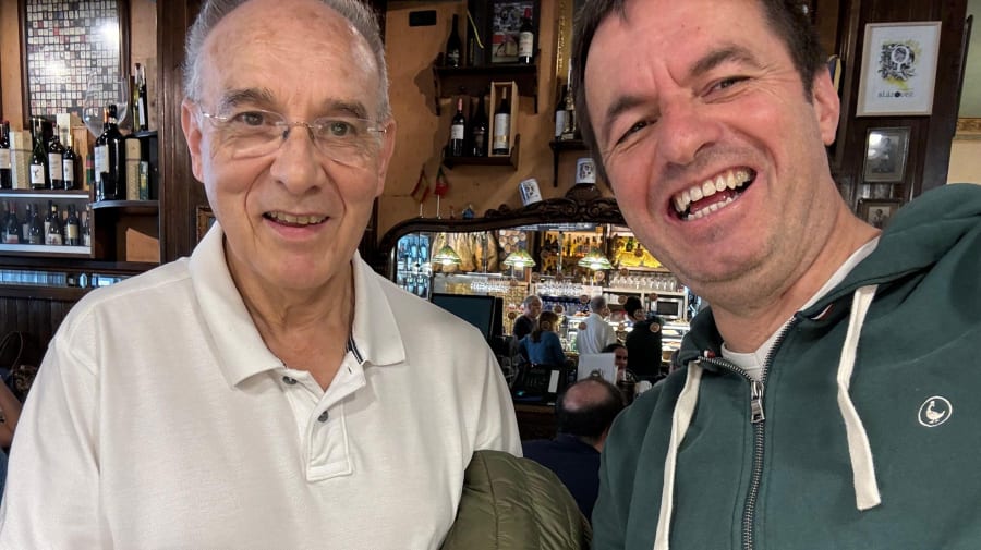Una fotografía que muestra a dos hombres sonrientes en un entorno de bar o restaurante, con una estantería de botellas al fondo.