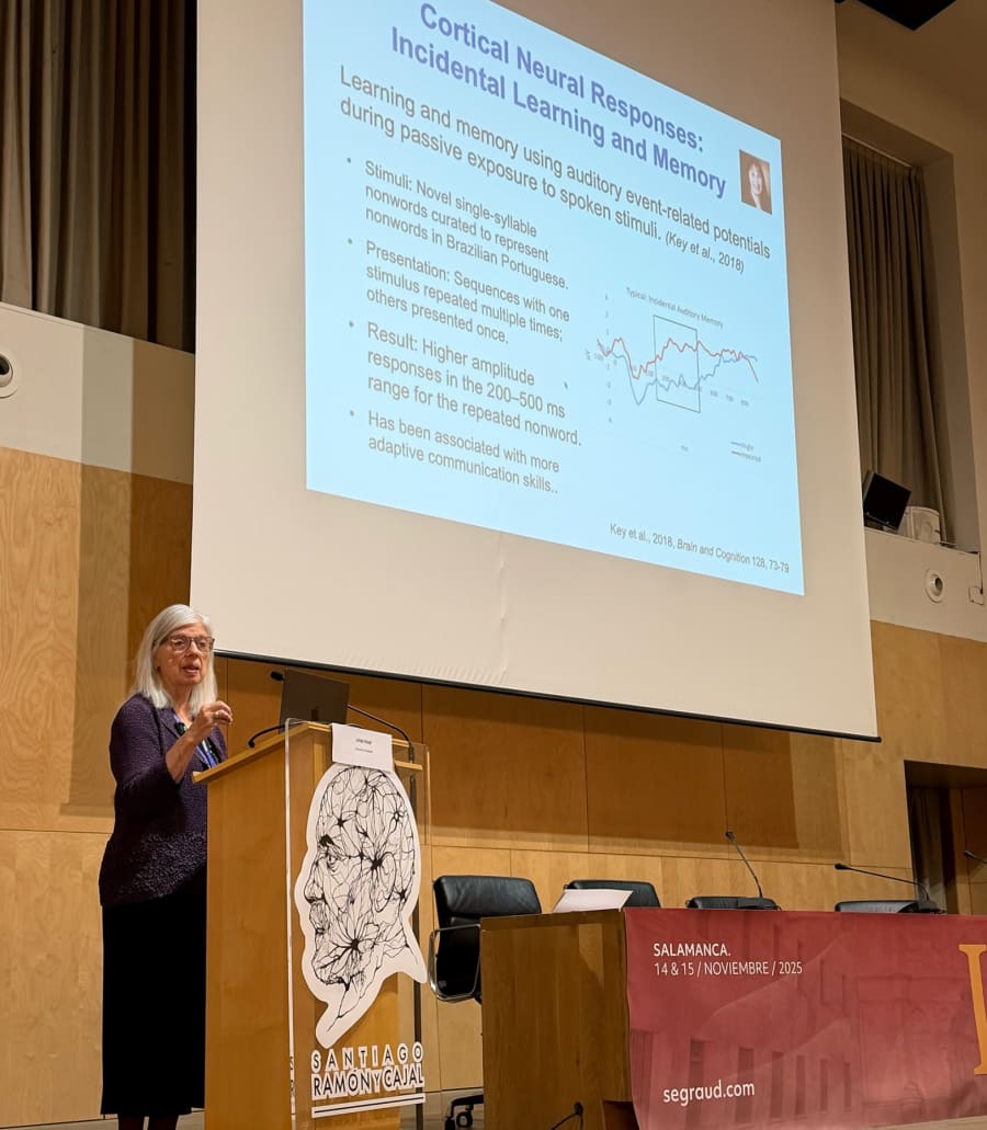 La profesora presenta en un congreso sobre auditiva, con una diapositiva que muestra gráficos y resultados relacionados con el aprendizaje incidental utilizando estímulos auditivos.
