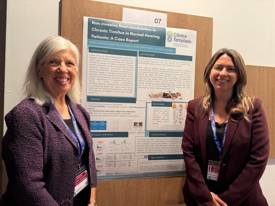 Dos mujeres sonrientes junto a un cartel de investigación sobre neuromodulación no invasiva en pacientes con tinnitus crónico en un congreso de audiología.