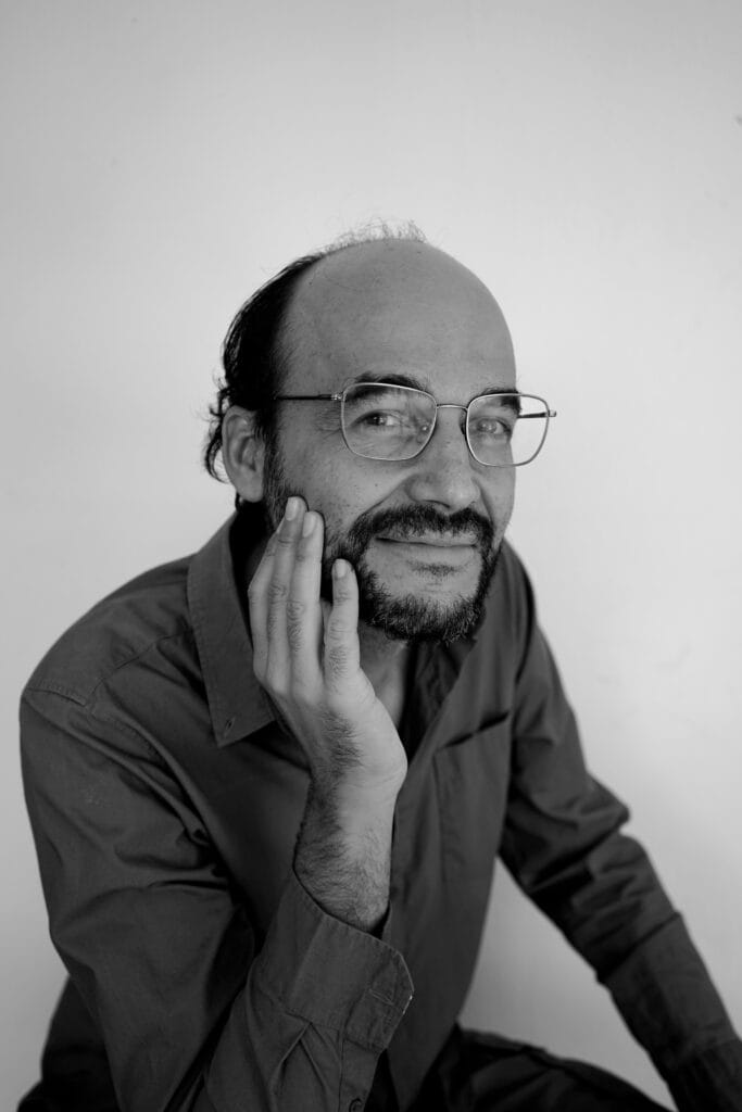 Retrato en blanco y negro de un hombre con gafas, sonriendo mientras sostiene su mano en la cara, vestido con una camisa de manga larga.
