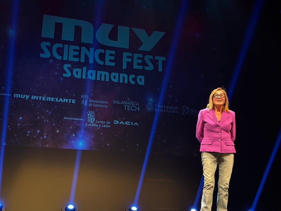 Presentadora en el escenario del MUY Science Fest Salamanca, con un fondo colorido que incluye el logotipo del evento y patrocinadores.