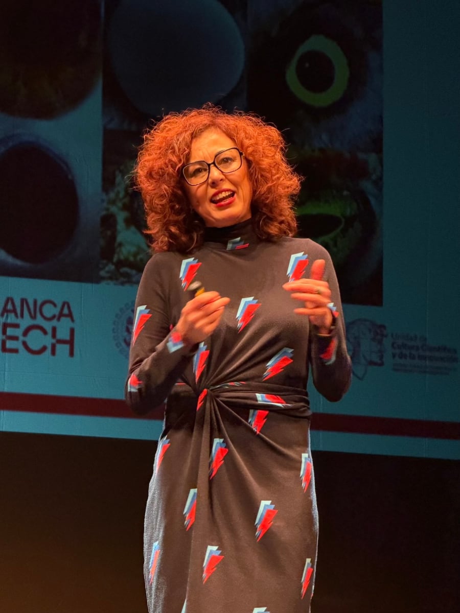 Mujer con cabello rizado y vestido de estampado colorido, hablando en un escenario, mientras presenta un tema relacionado con la ciencia.