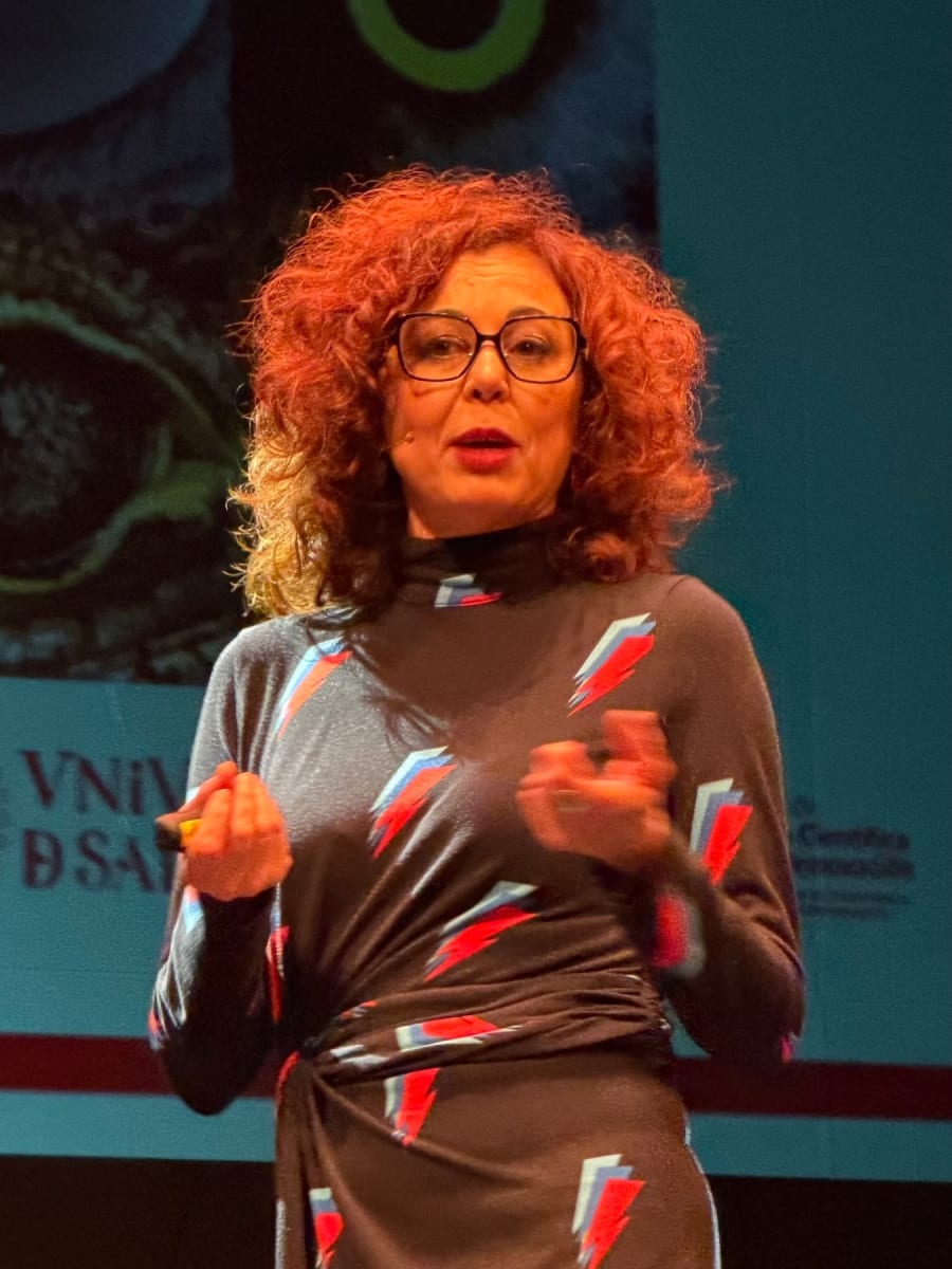 Mujer presentando en un escenario durante el festival de ciencia, con un vestido de patrones coloridos y expresión entusiasta.