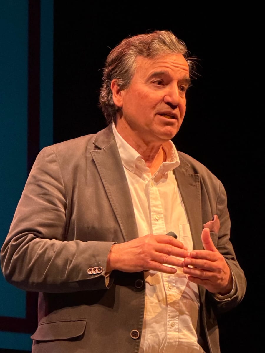 Hombre de pie hablando en un escenario, con un fondo oscuro, vistiendo un blazer gris y una camisa blanca, gesticulando con las manos.