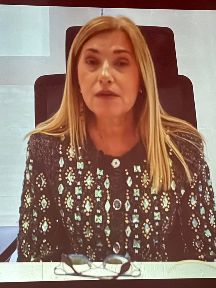 Una mujer con cabello largo y rubio, vestida con una chaqueta decorativa y un fondo gris, habla frente a una cámara.