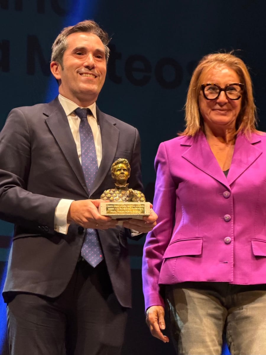 Dos personas están en un escenario durante un evento. Un hombre lleva un traje oscuro y sostiene un premio en forma de figura dorada, mientras que una mujer a su lado viste una chaqueta rosa y sonríe. Al fondo se puede distinguir un panel con texto en letras grandes.