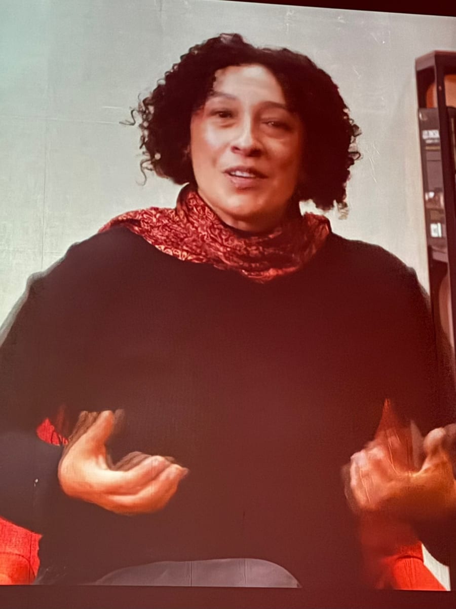 Una mujer con cabello rizado y una bufanda roja, dando una charla o presentación, mientras sonríe y gesticula con las manos.