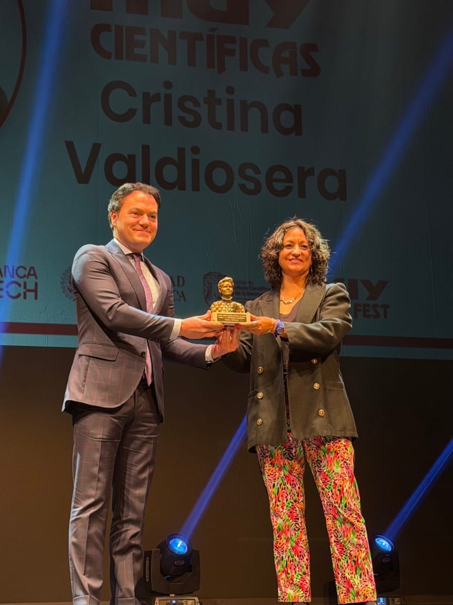 Una mujer recibiendo un premio de manos de un hombre en un escenario iluminado, con un fondo que incluye el texto