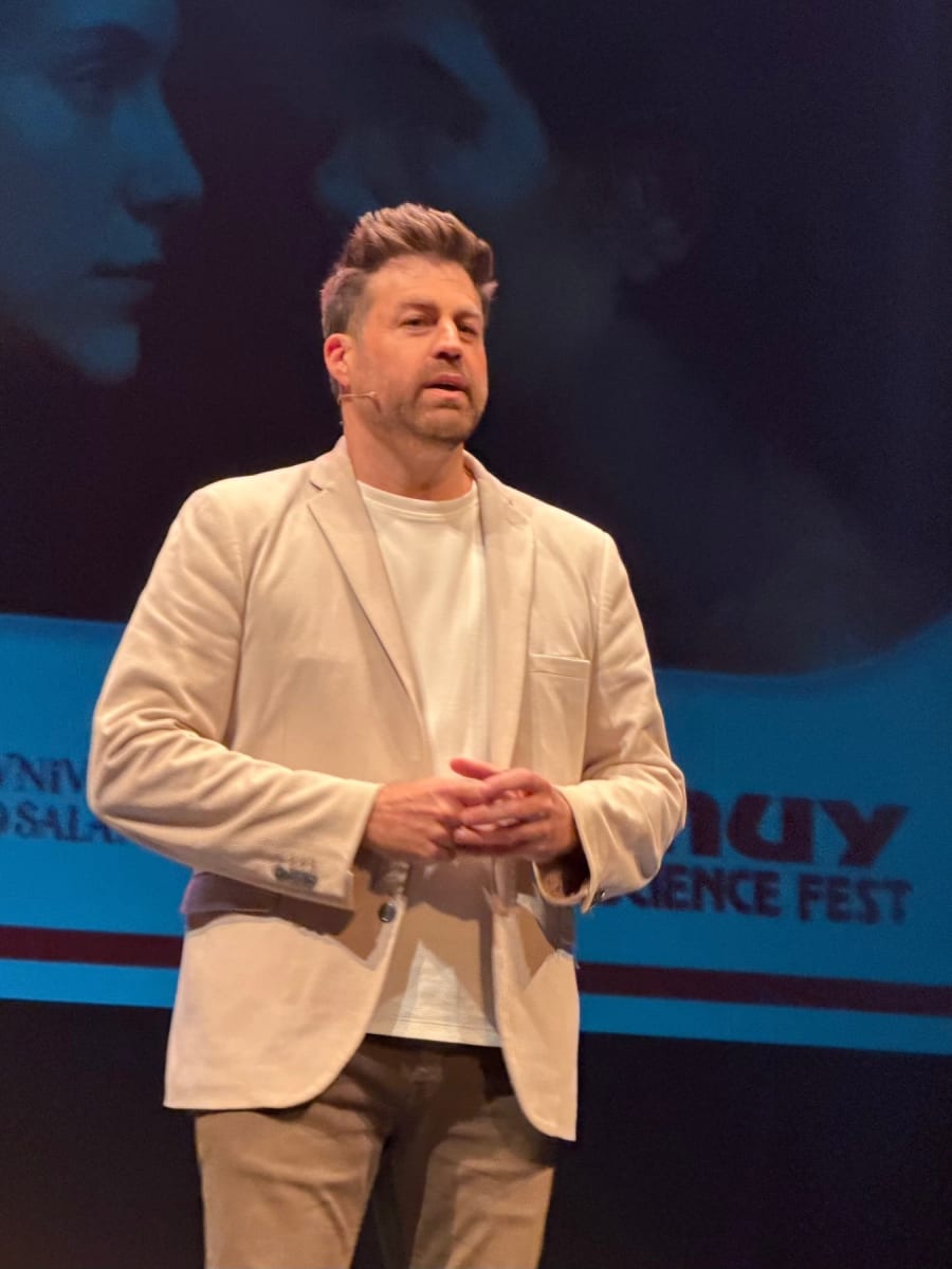 Un hombre de pie en un escenario del Teatro Liceo durante el MUY Science Fest 2025, vestido con un abrigo claro y una camiseta blanca, mientras se prepara para hablar ante la audiencia.