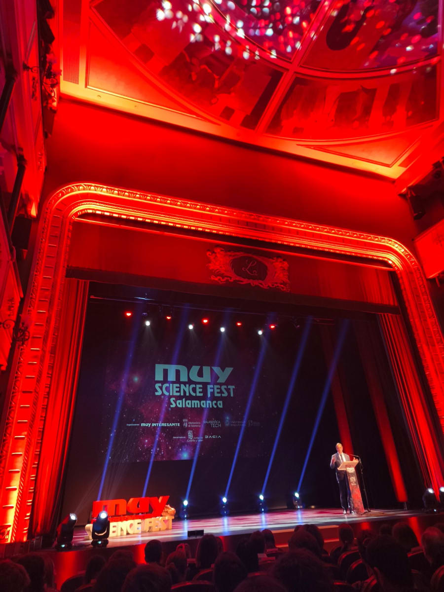 Escenario del MUY Science Fest en Salamanca, con iluminación roja y un presentador hablando detrás de un micrófono, mientras el público observa desde sus asientos.