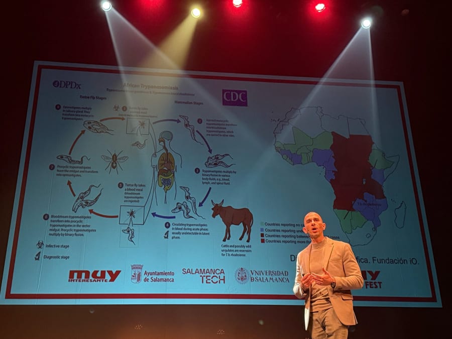 Un ponente presenta un diagrama sobre la enfermedad del sueño africana, ilustrando el ciclo de vida de la mosca tsetsé y su relación con los mamíferos en el MUY Science Fest 2025 en el Teatro Liceo de Salamanca.