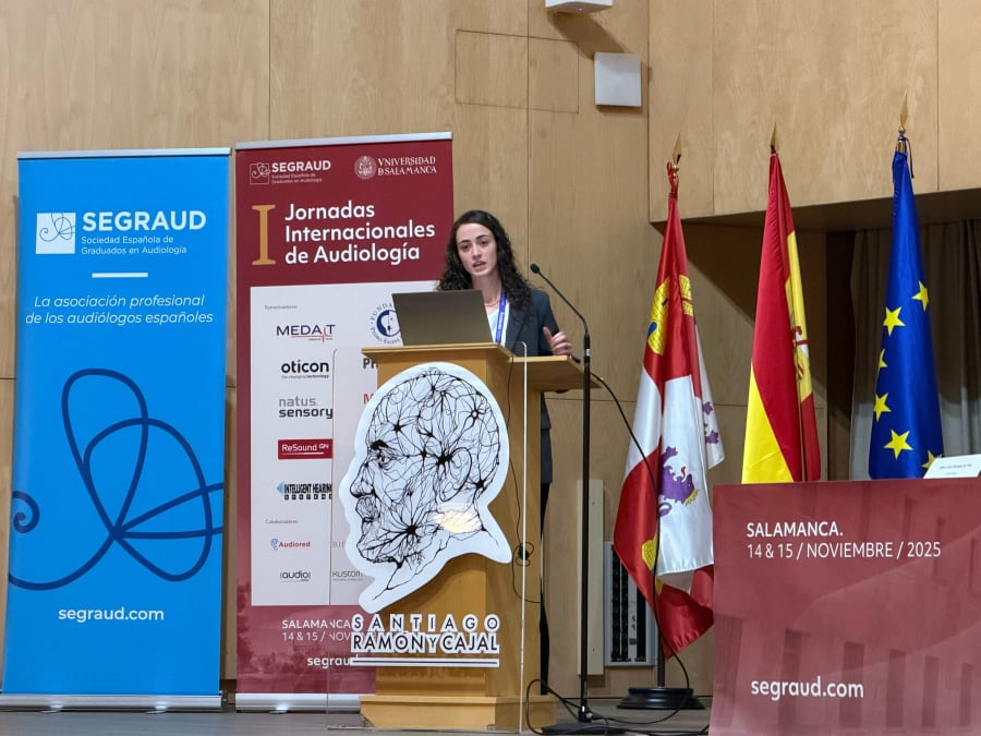 Presentadora en una conferencia sobre audiología, con banderas de España y la UE en el fondo, durante las I Jornadas Internacionales de Audiología en Salamanca.