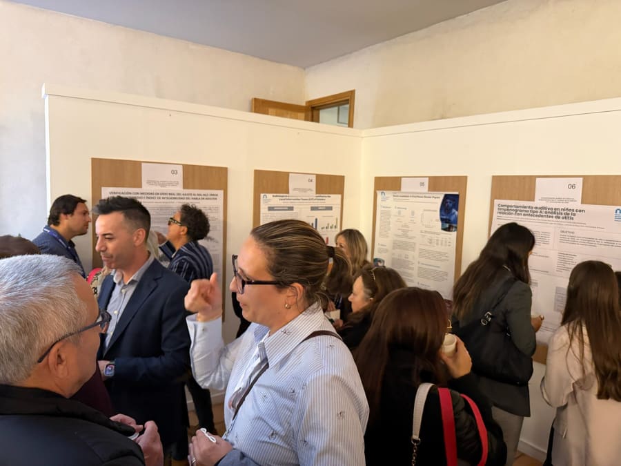 Un grupo de personas interactuando en un evento académico, rodeados de carteles de pósters de investigación sobre audiología. Varias personas están observando los pósters y discutiendo entre sí.