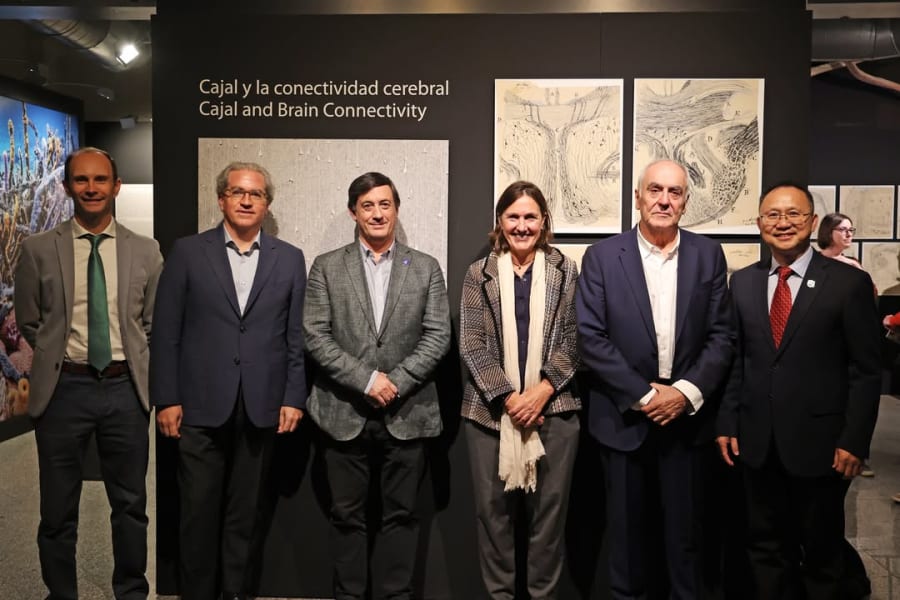 Un grupo de seis personas posando frente a un panel que presenta la exposición "Cajal y la conectividad cerebral". Al fondo se aprecian obras gráficas relacionadas con el tema de la neurociencia.