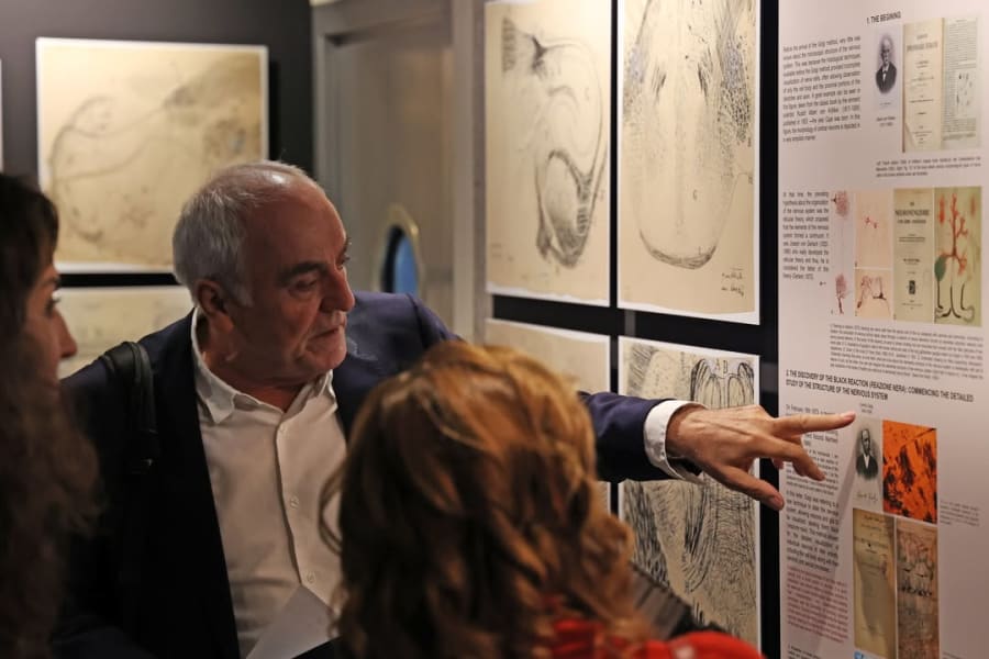 Un hombre de pie en una sala de exposiciones, señalando un panel informativo, mientras explica a dos personas sobre ilustraciones científicas en la pared detrás de ellos, que muestra obras relacionadas con la neurociencia.