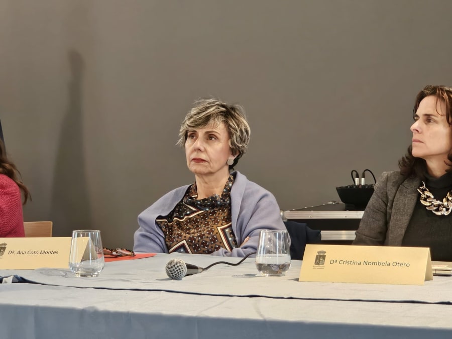 Tres mujeres sentadas en una mesa durante un evento académico, con un micrófono y copas de agua sobre la mesa.