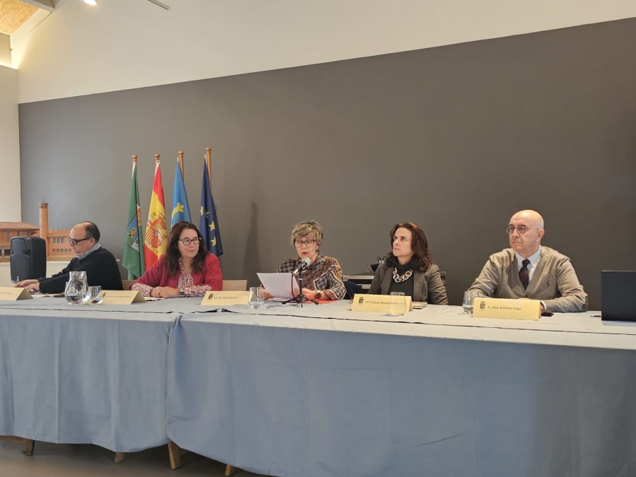 Mesa redonda en el Museo de la Mina de Arnao con cuatro ponentes, quienes discuten sobre la ciencia y el legado de Santiago Ramón y Cajal, con banderas de España y regiones visibles al fondo.