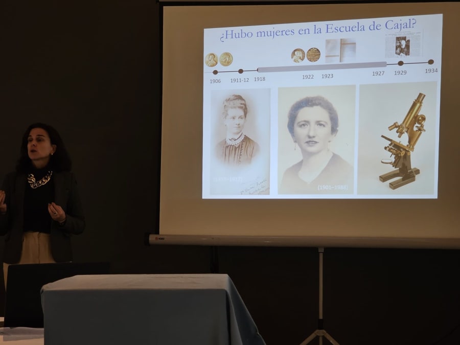 Una presentadora habla sobre las mujeres en la Escuela de Cajal, junto a una presentación que incluye fotos de científicas y un microscopio.