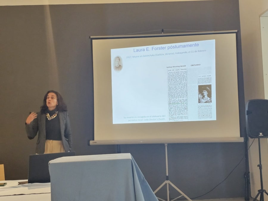 Ponente en una conferencia, hablando sobre Laura E. Forster, con una presentación en pantalla que incluye detalles sobre su vida y un obituario.