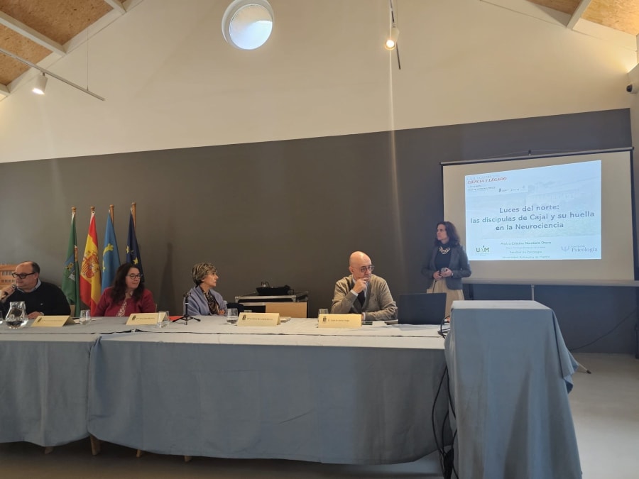 Ponente presentando en la jornada "Luces del norte: las discípulas de Cajal y su huella en la Neurociencia", con mesa de conferenciantes y banderas al fondo.