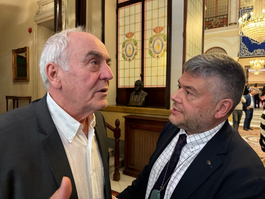 Dos hombres conversando en un interior elegante, con detalles arquitectónicos visibles en el fondo. Uno de ellos lleva un traje oscuro y el otro una camisa blanca debajo de un saco. La escena refleja un ambiente académico o de evento formal.