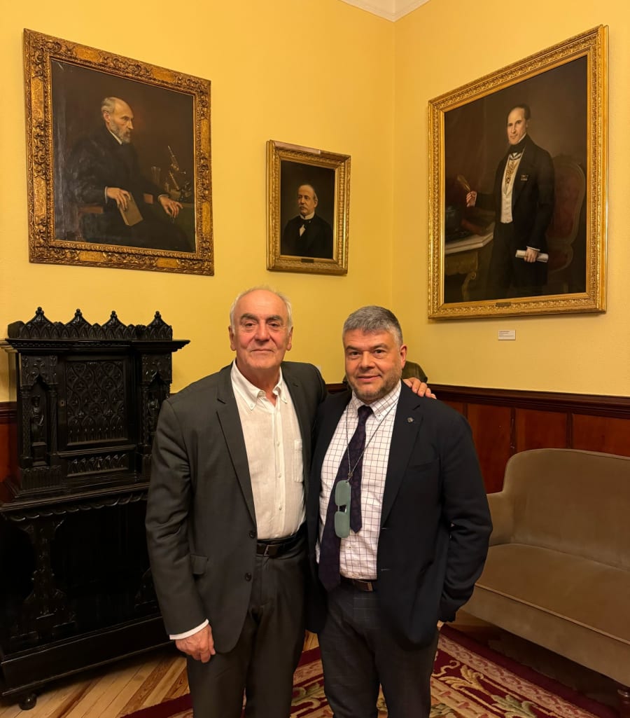 Dos hombres de pie frente a un mueble en una sala con paredes amarillas, retratos de figuras históricas enmarcados en las paredes, y un sofá beige al fondo.