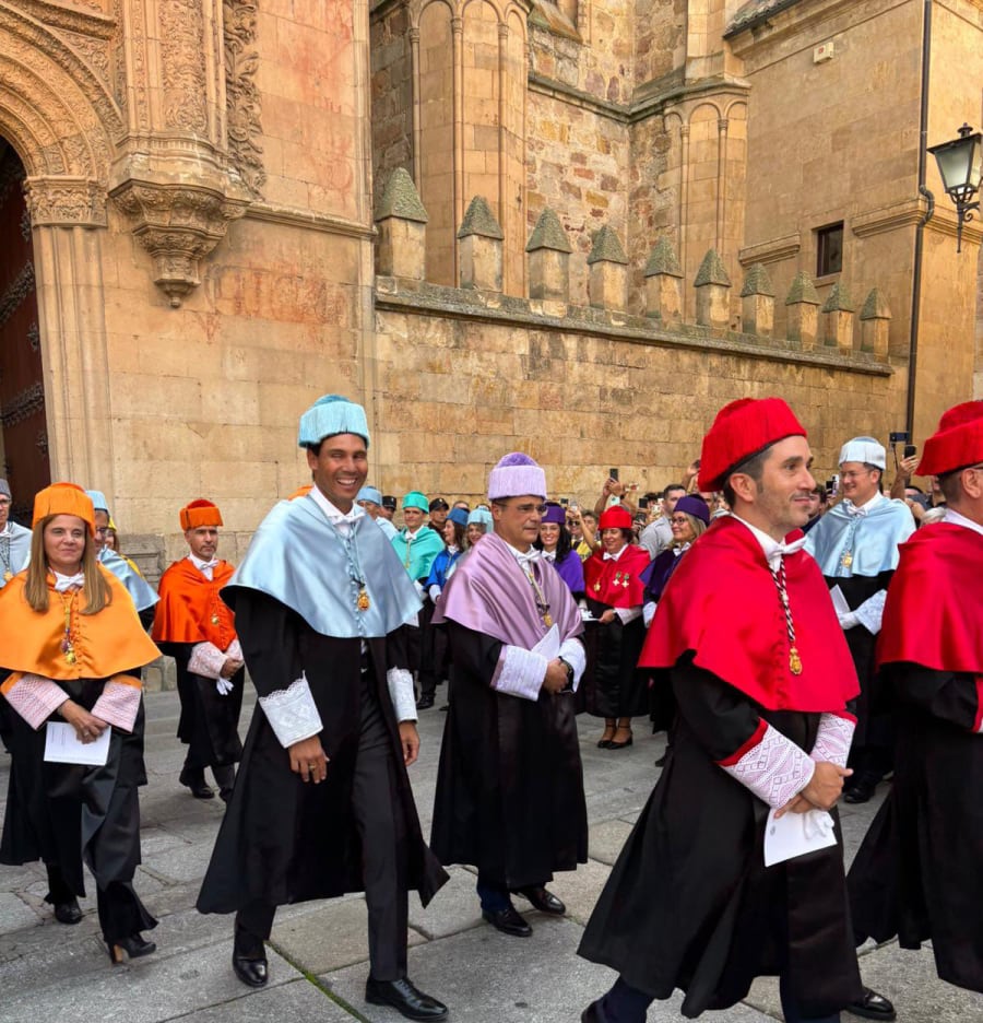 Un grupo de académicos en trajes ceremoniales coloridos, caminando en el exterior del Paraninfo de la Universidad de Salamanca, con una multitud al fondo observando el evento.
