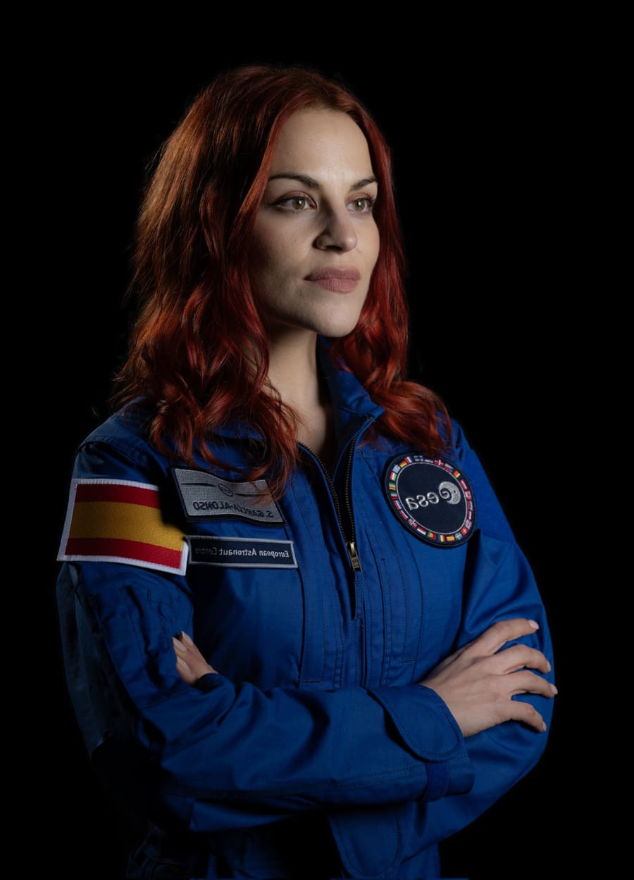 Retrato de una mujer con traje de astronauta azul, destacando la insignia de la Agencia Espacial Europea y la bandera de España, con cabello largo y rojo, mirándose al frente en un fondo negro.