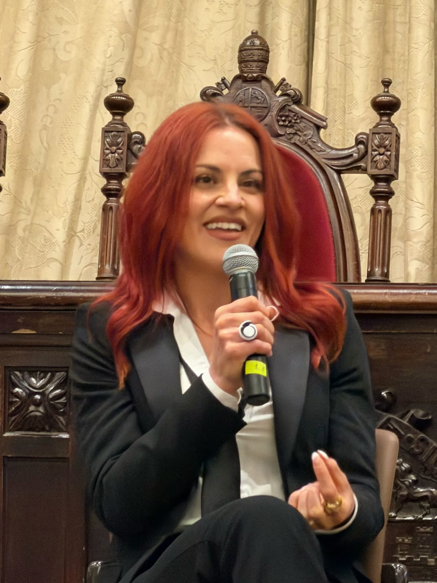 Mujer con cabello largo y rojo, vistiendo un traje negro y una blusa blanca, hablando en un micrófono en un evento, con un fondo de cortinas y sillas decorativas.