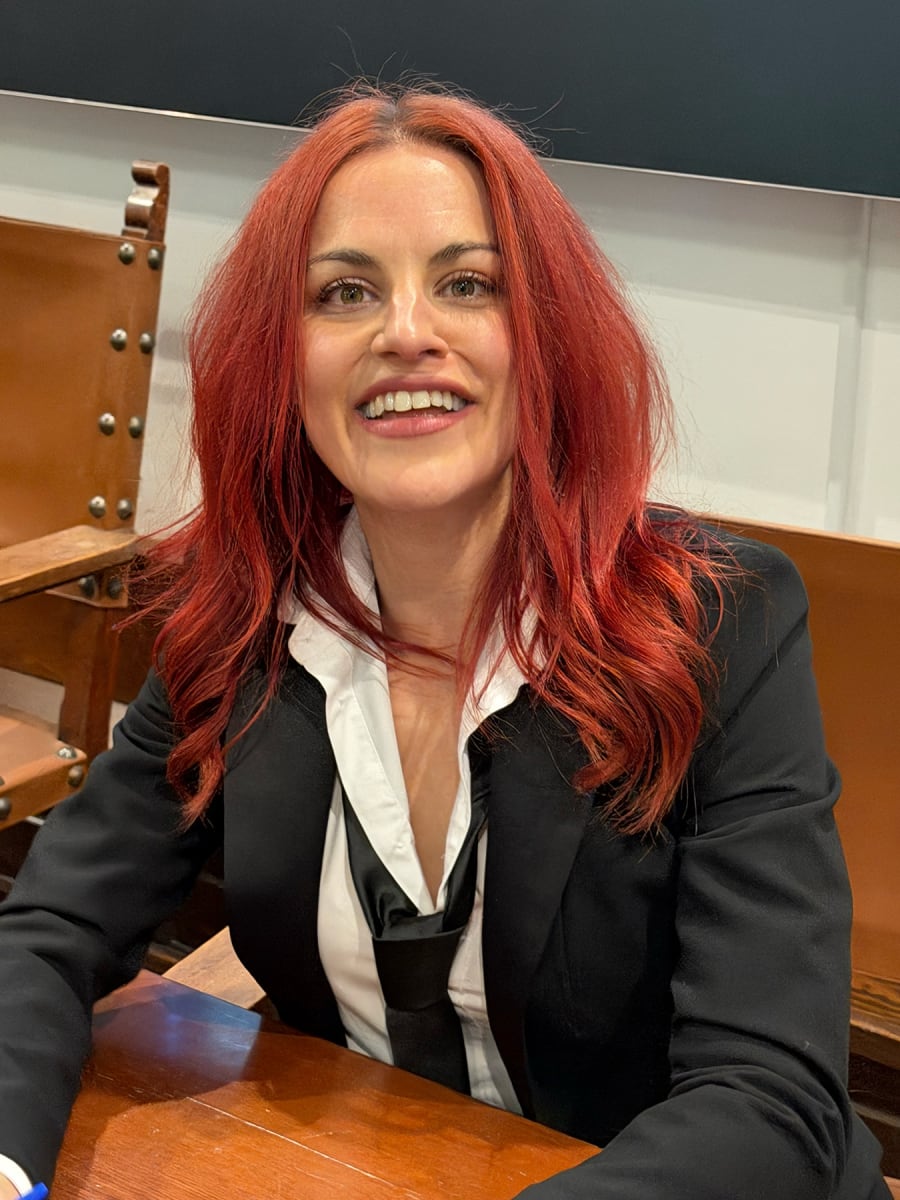 Retrato de una mujer con cabello rojo, sonriendo mientras viste un traje negro y camisa blanca, sentada en una mesa de madera.