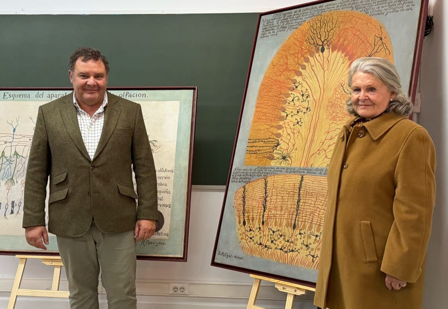Un hombre y una mujer posan juntos frente a dos obras de arte que representan diagramas del sistema olfativo y neuronal, en un entorno educativo.
