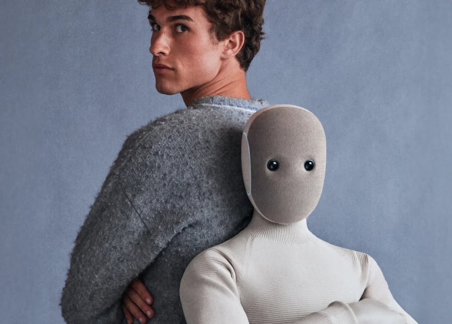 Un modelo masculino posando de espaldas con un muñeco de textura suave que parece humano, ambos frente a un fondo gris.