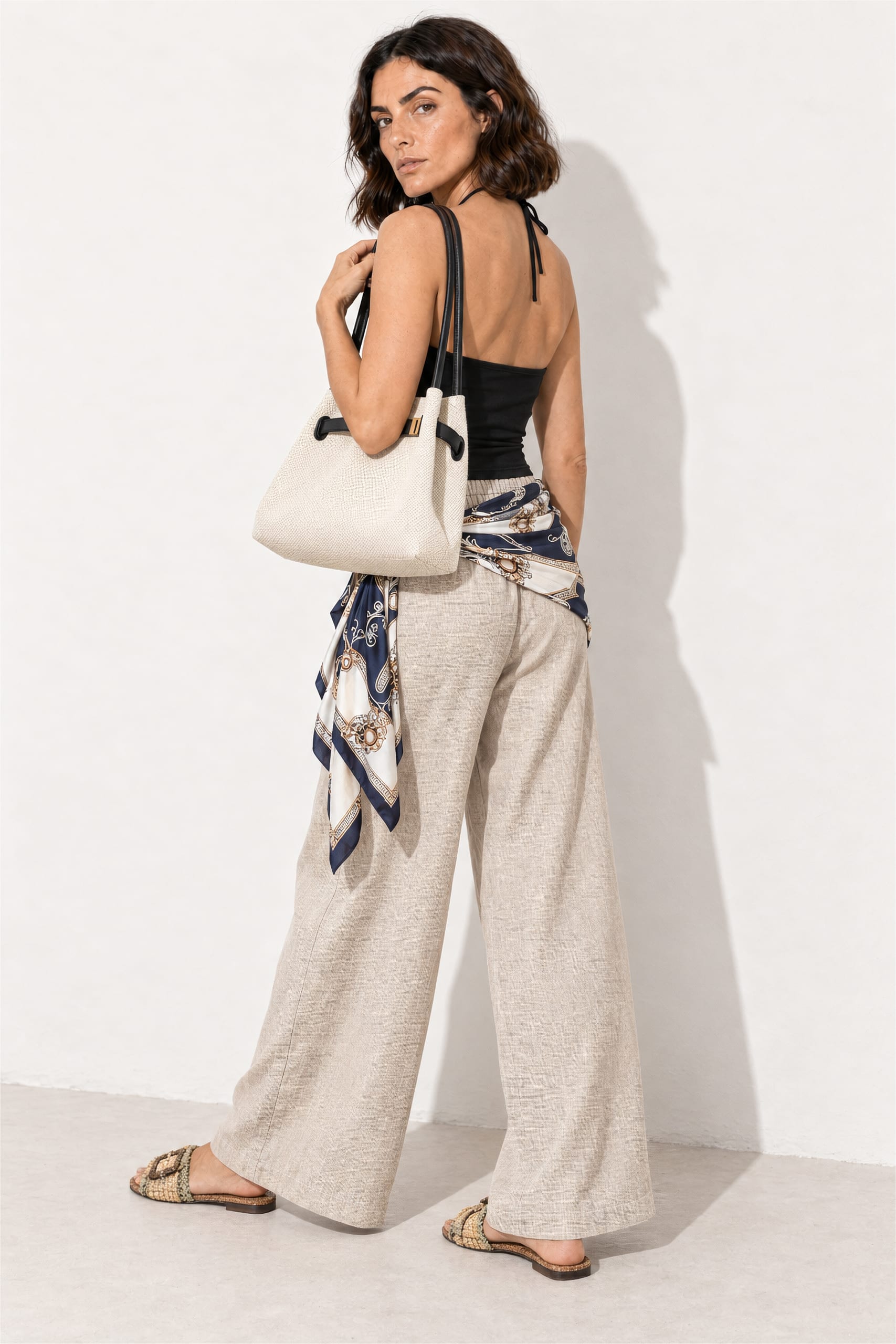 Back view of a navy silk scarf wrapped around cream linen-blend palazzo pants and tied at the side with a long tail, styled with a black halter top and structured tote.