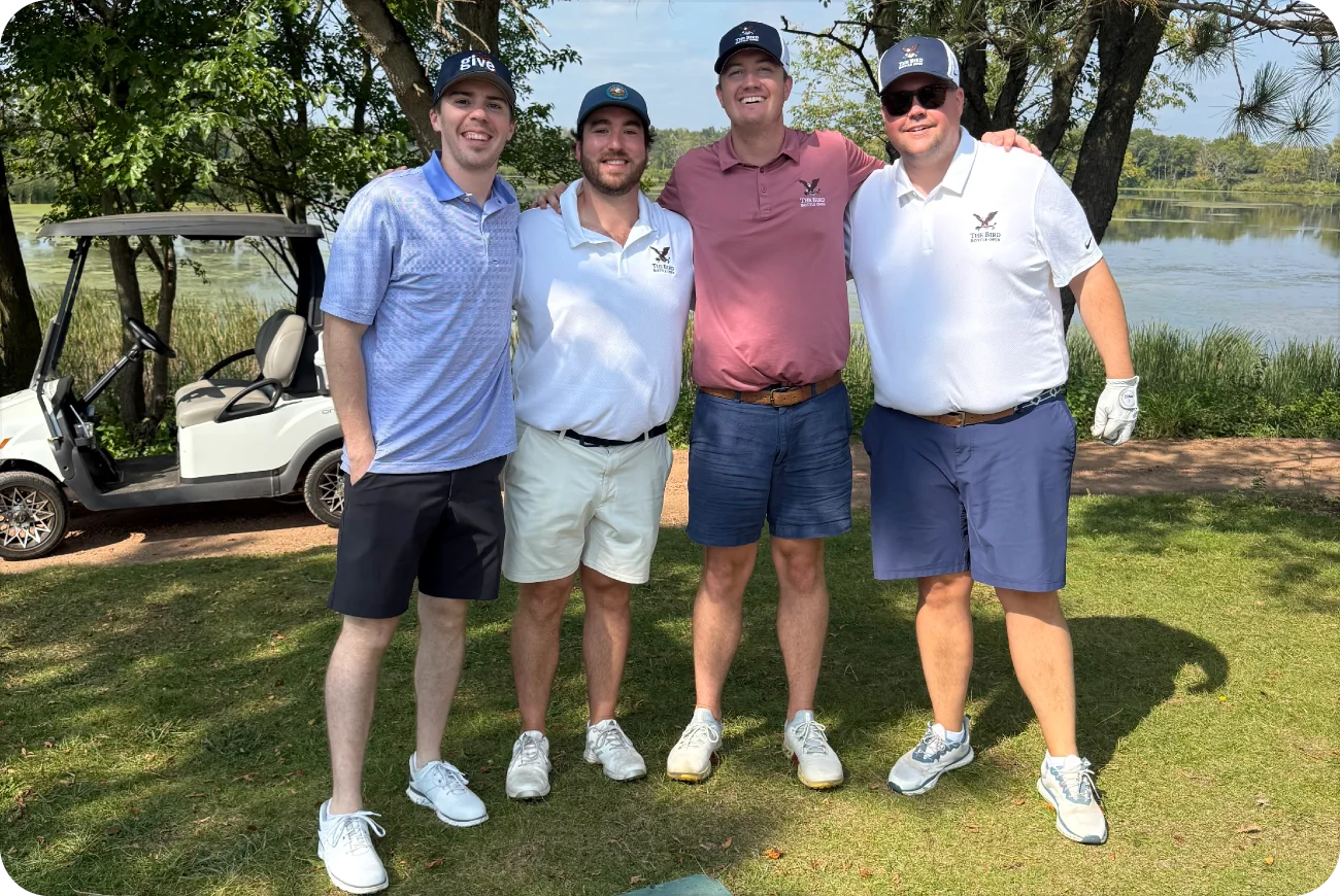 Four men posing together on a golf course with a cart and pond in the background