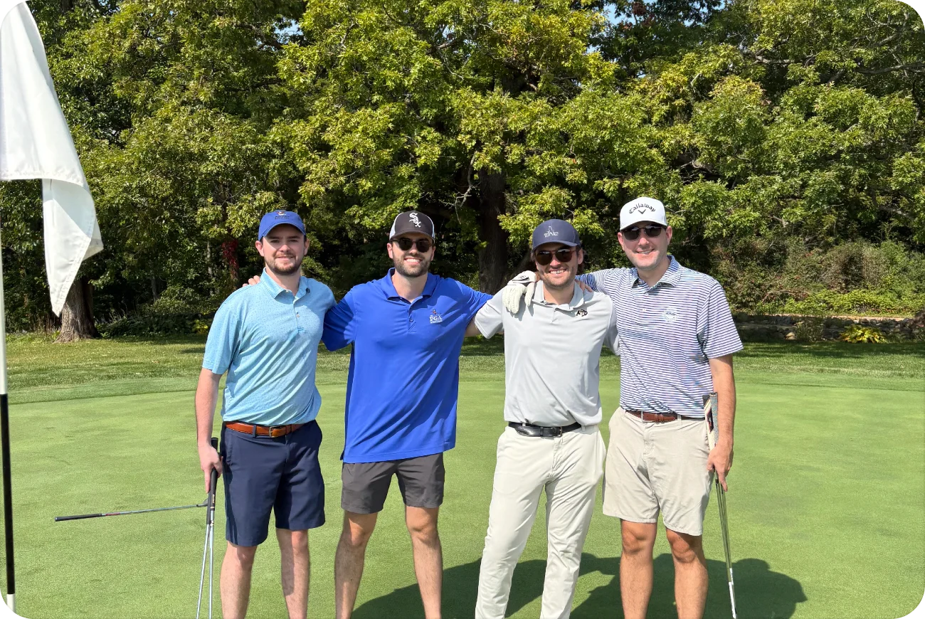 Four men in golf attire posing on a green near a flag