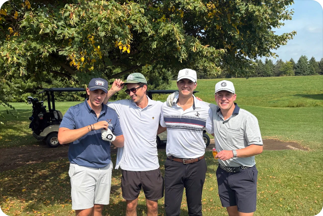 Four men smiling together under a tree with a golf cart nearby