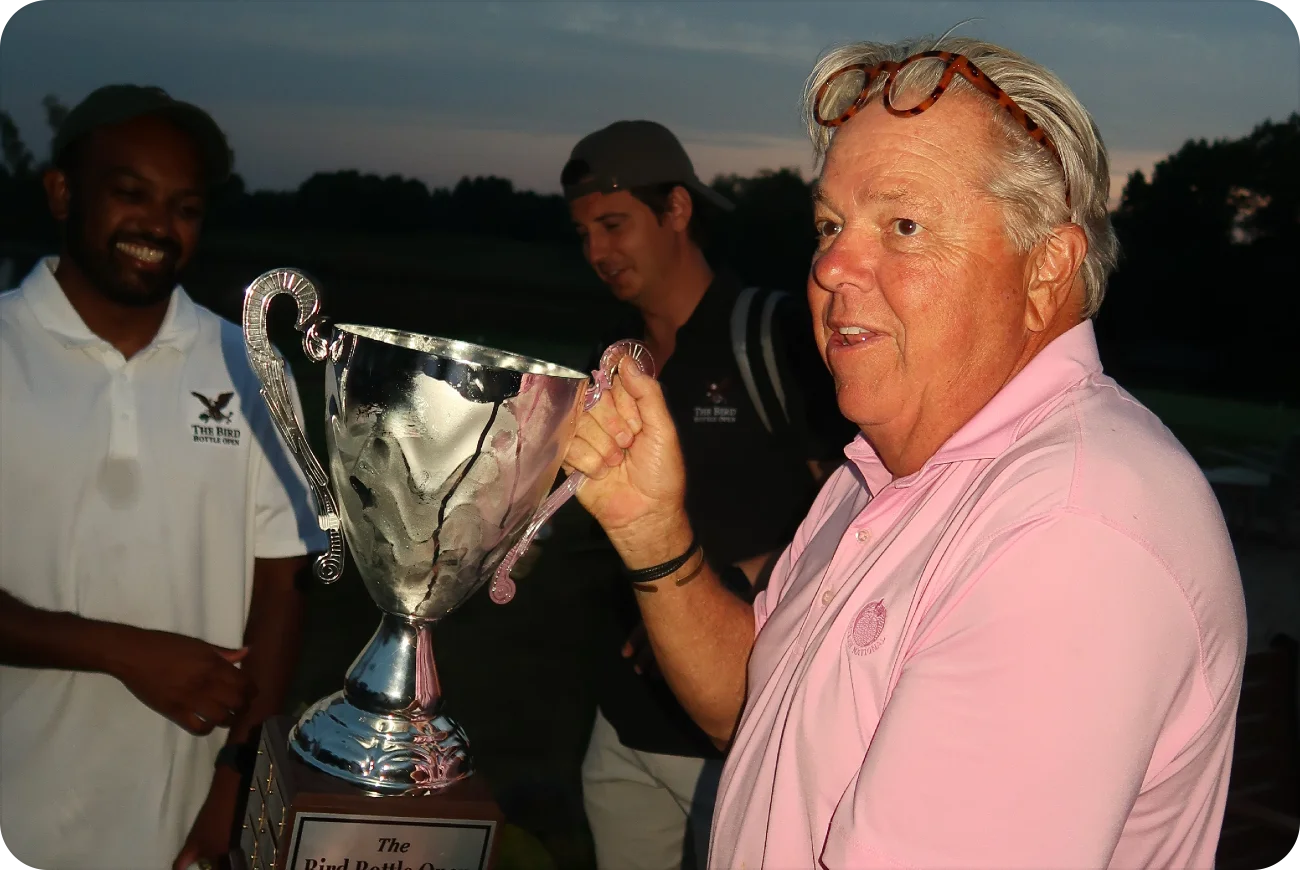 Man in pink polo hoisting the Bird Bottle Open championship trophy at dusk