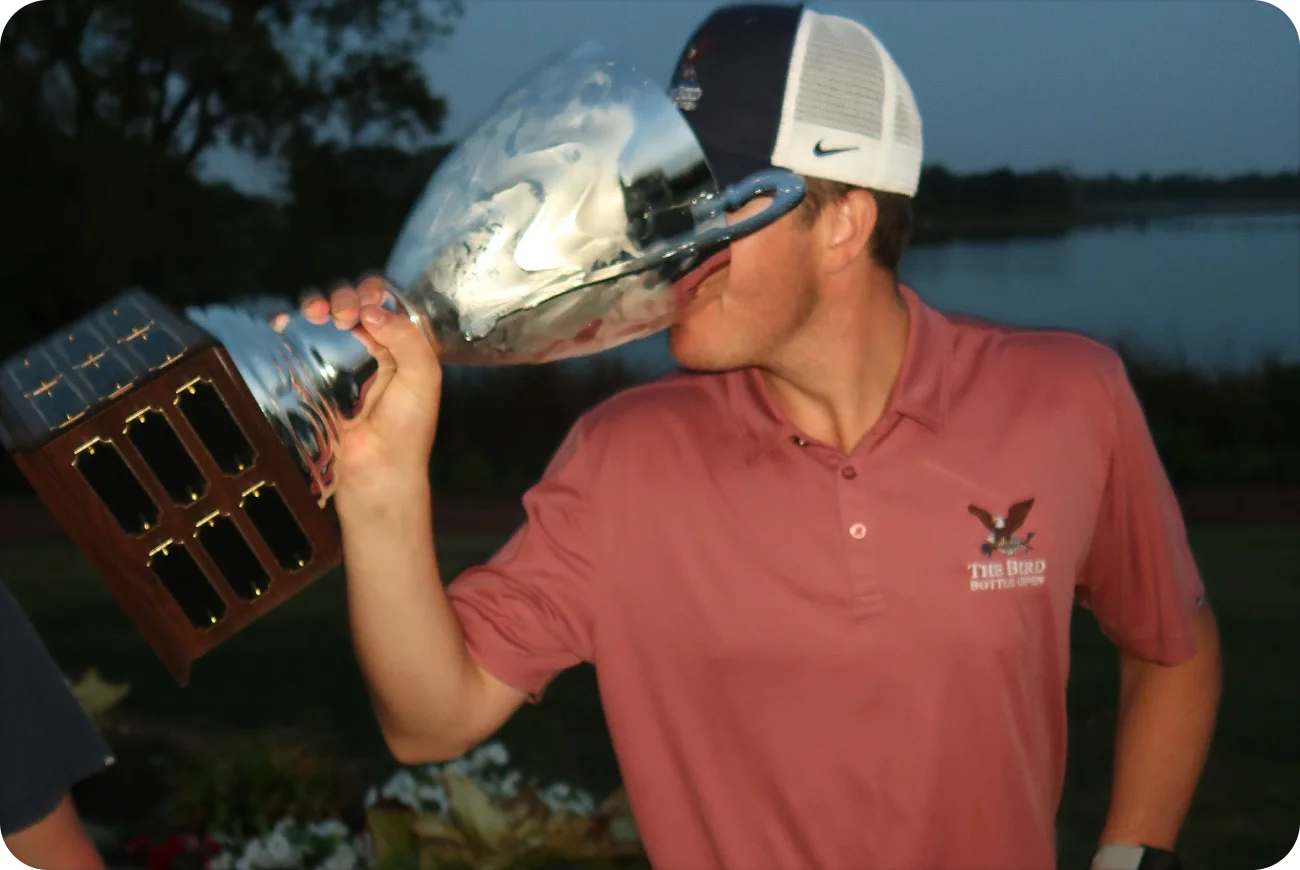 Man kissing the Bird Bottle Open championship trophy at dusk by the water