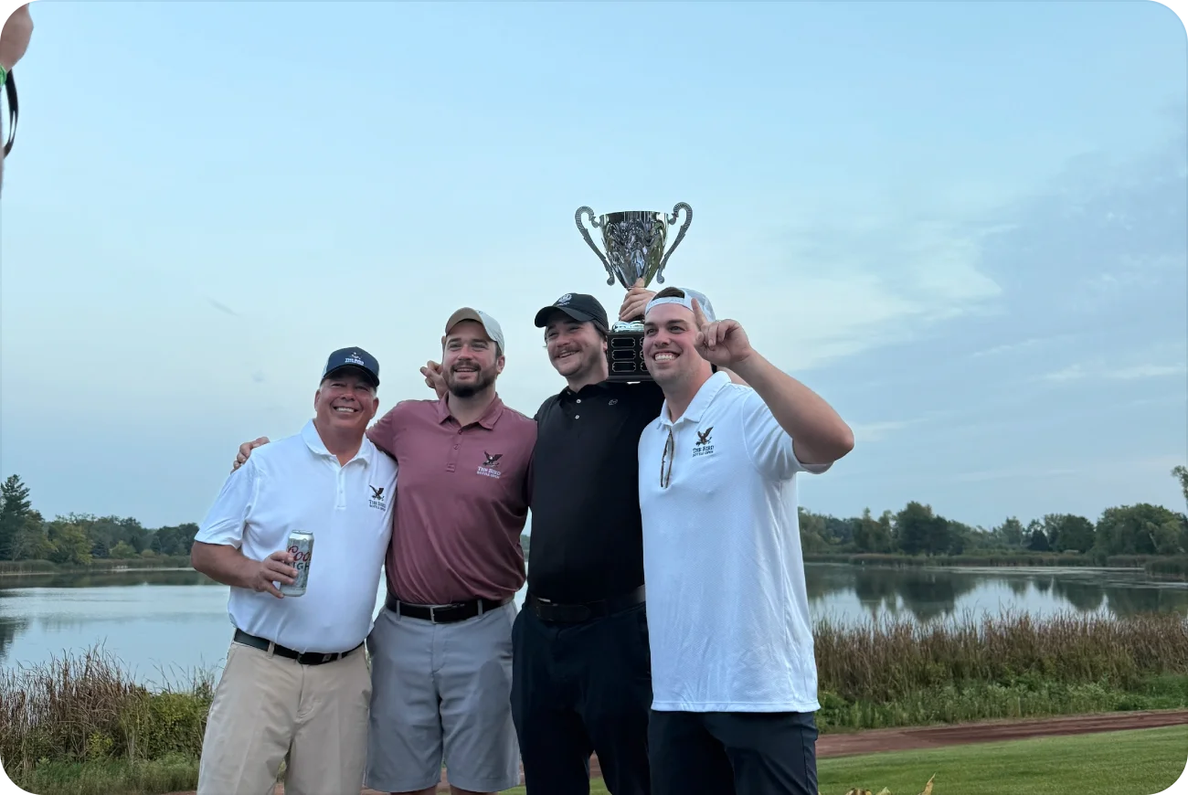 Four men celebrating with the Bird Bottle Open trophy raised by a lake at dusk