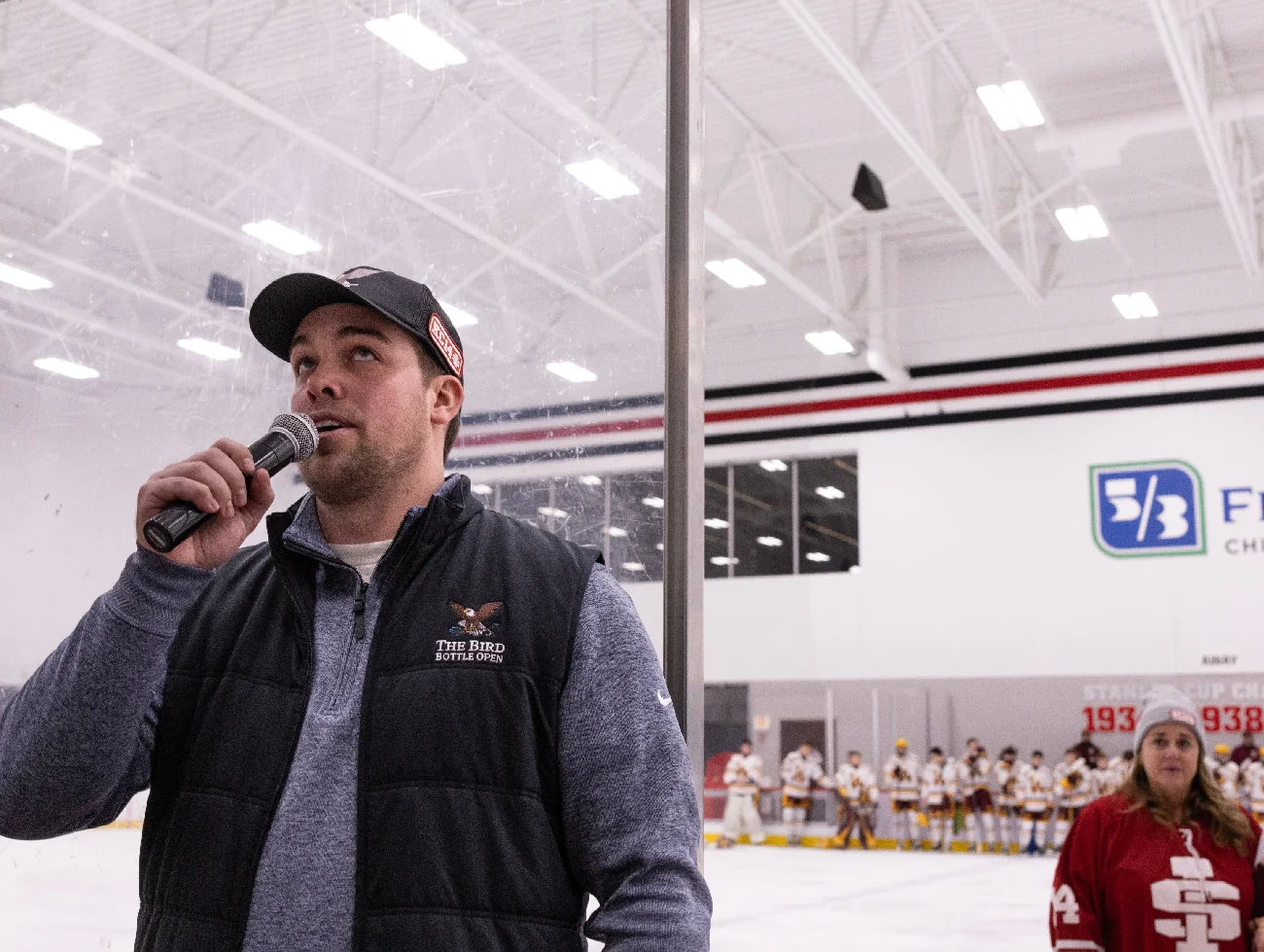 Man speaking into a microphone at the rink with both teams lined up on the ice