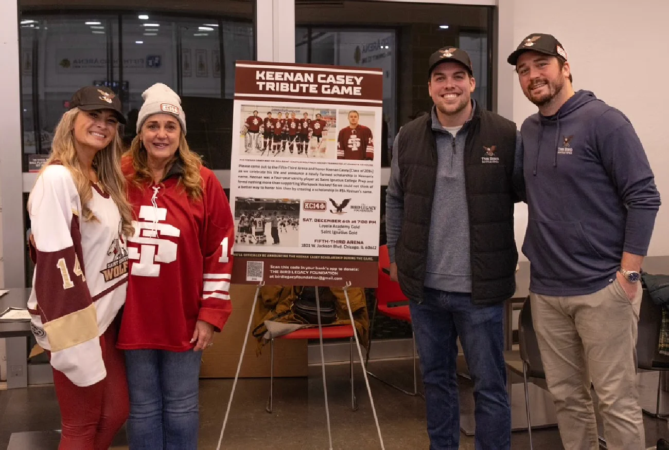 Four people posing next to the Keenan Casey Tribute Game event poster at the arena