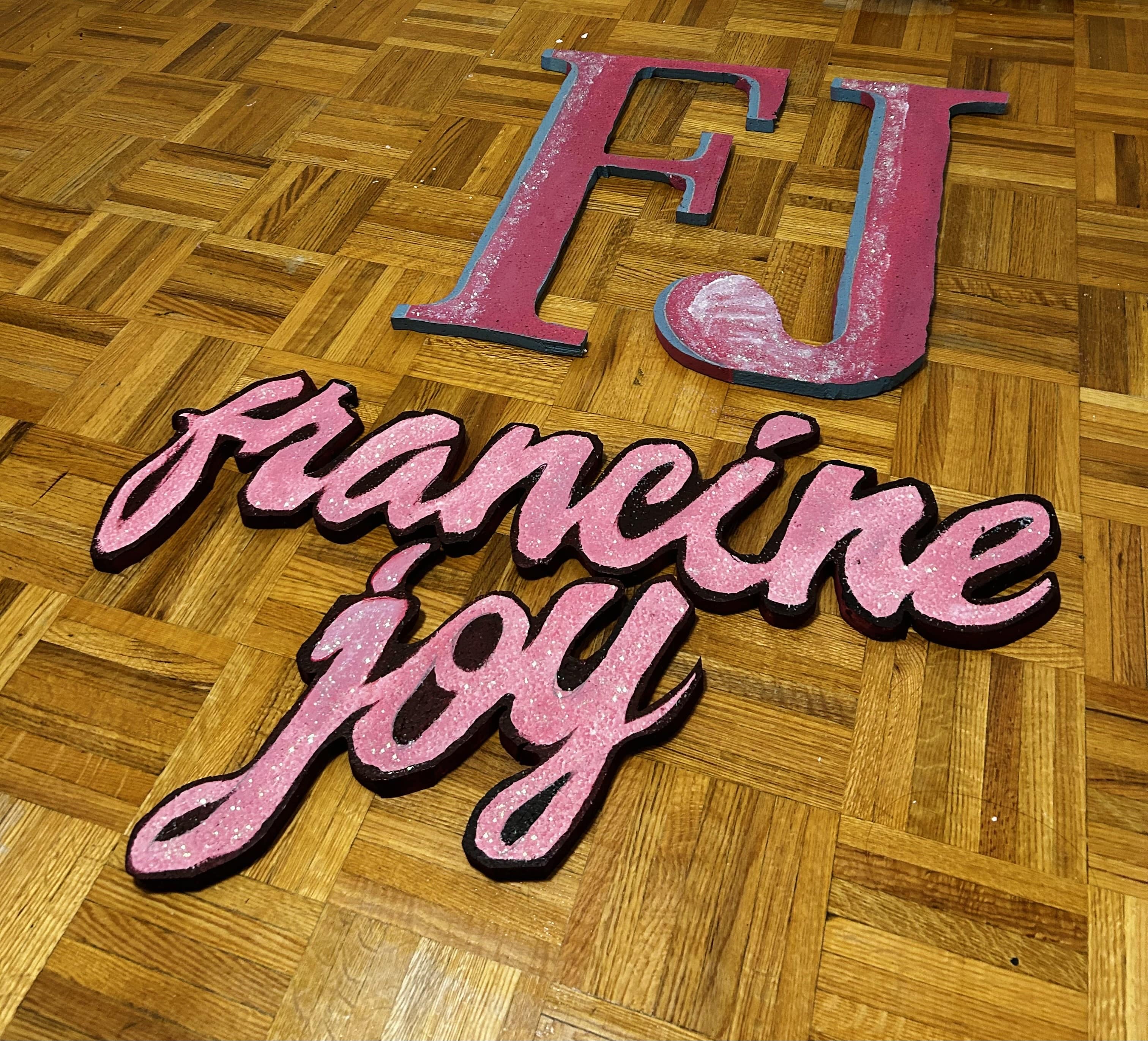 Pink painted styrofoam cutouts of large letters F and J, and a calligraphy cutout on a wooden plank floor.