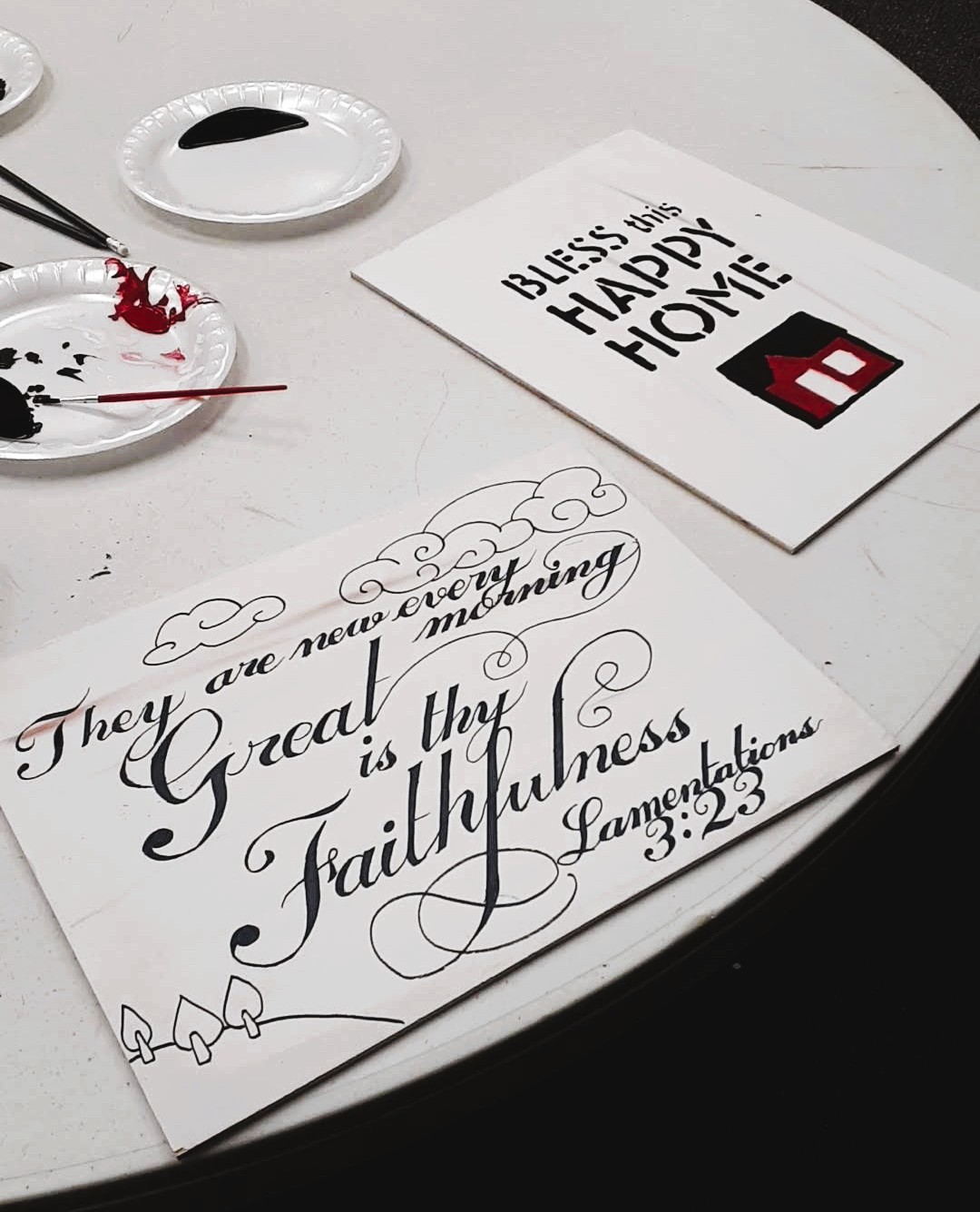 Two wooden canvases on a white round table, one with inscription 'Bless this Happy Home' and another with a calligraphy of Lamentations 3:23.