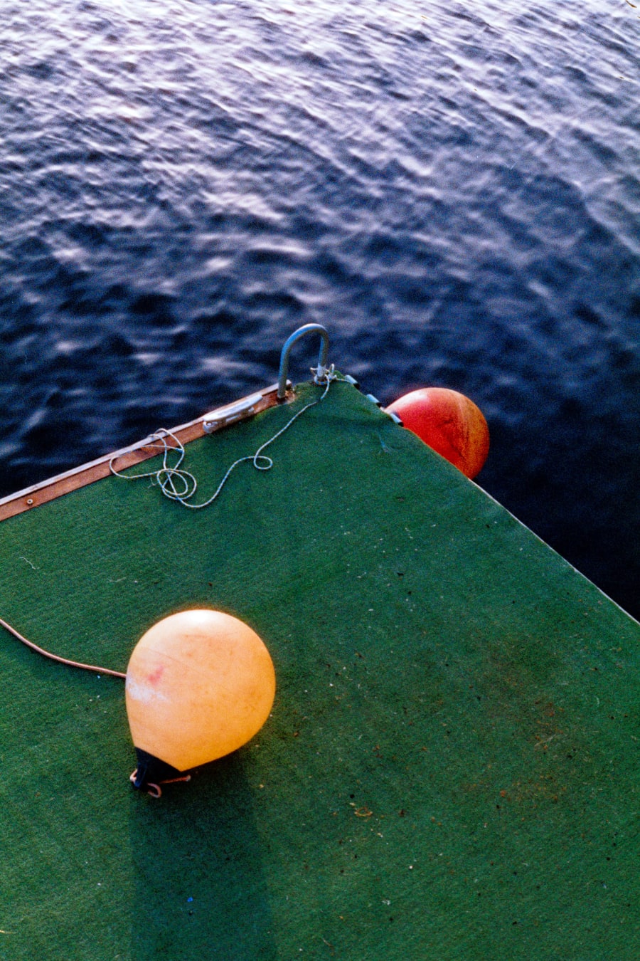 Colour photograph looking down at two weathered orange and yellow buoys resting on the green deck of a moored boat, with dark rippled water beyond.