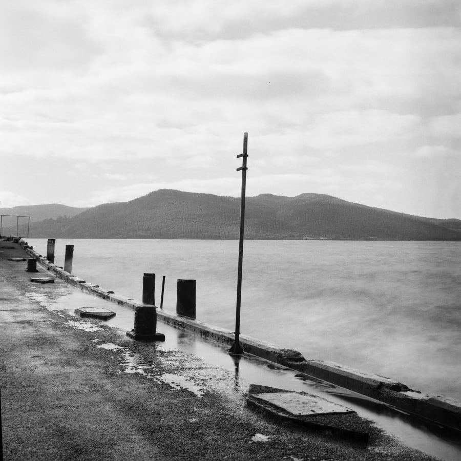 Black and white long exposure of a rain-slicked jetty lined with mooring bollards, still water smoothed flat, and forested hills across the bay under heavy cloud.