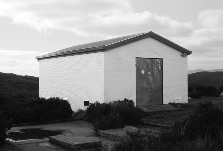 Black and white photograph of a small white brick shed with a corrugated roof and rusted steel door, standing alone in low coastal scrub with hills behind.