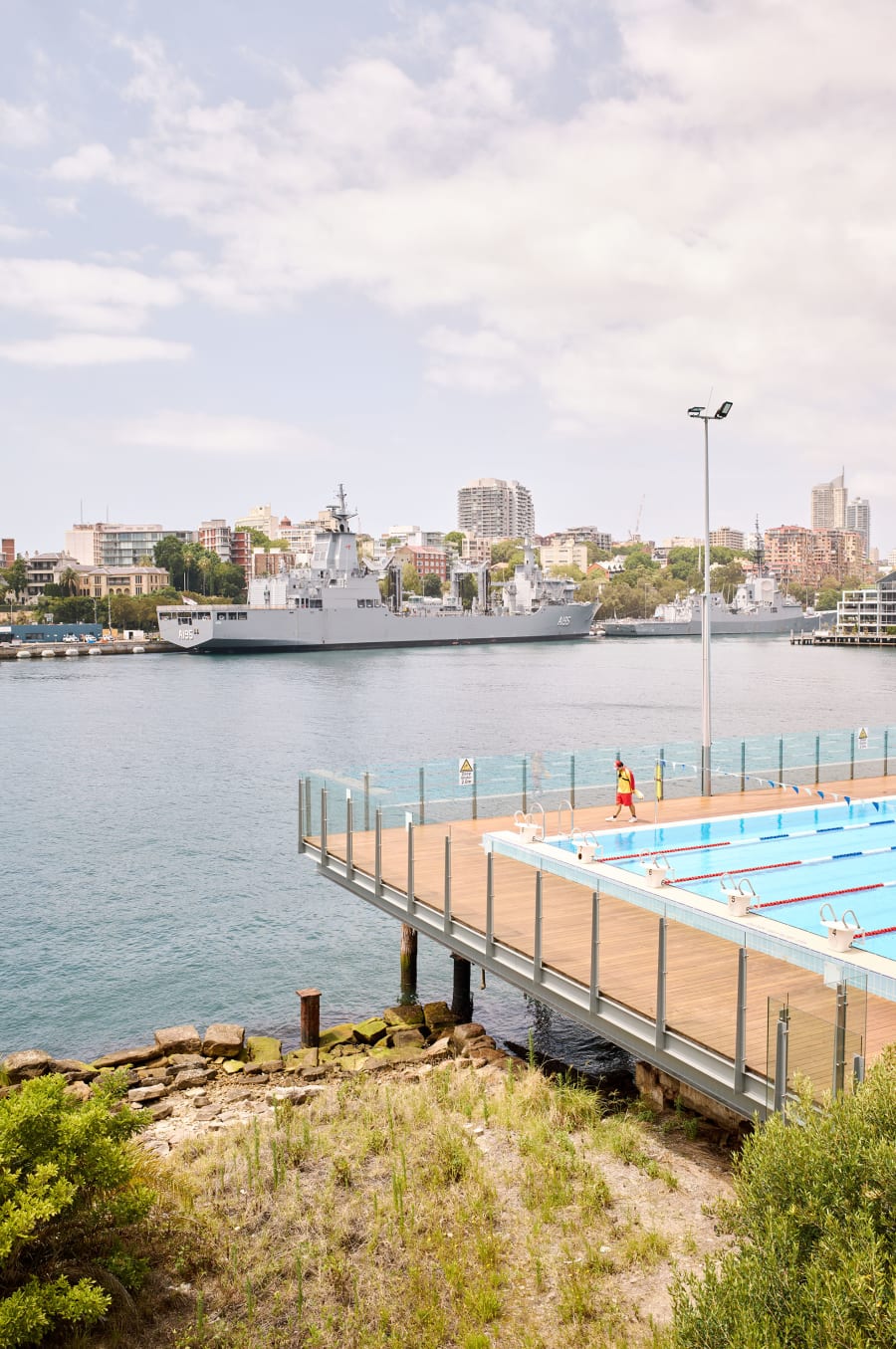 Colour photograph of Sydney Harbour: navy frigates moored beneath the city skyline, with a lifeguard walking the deck of a harbourside tidal swimming pool.