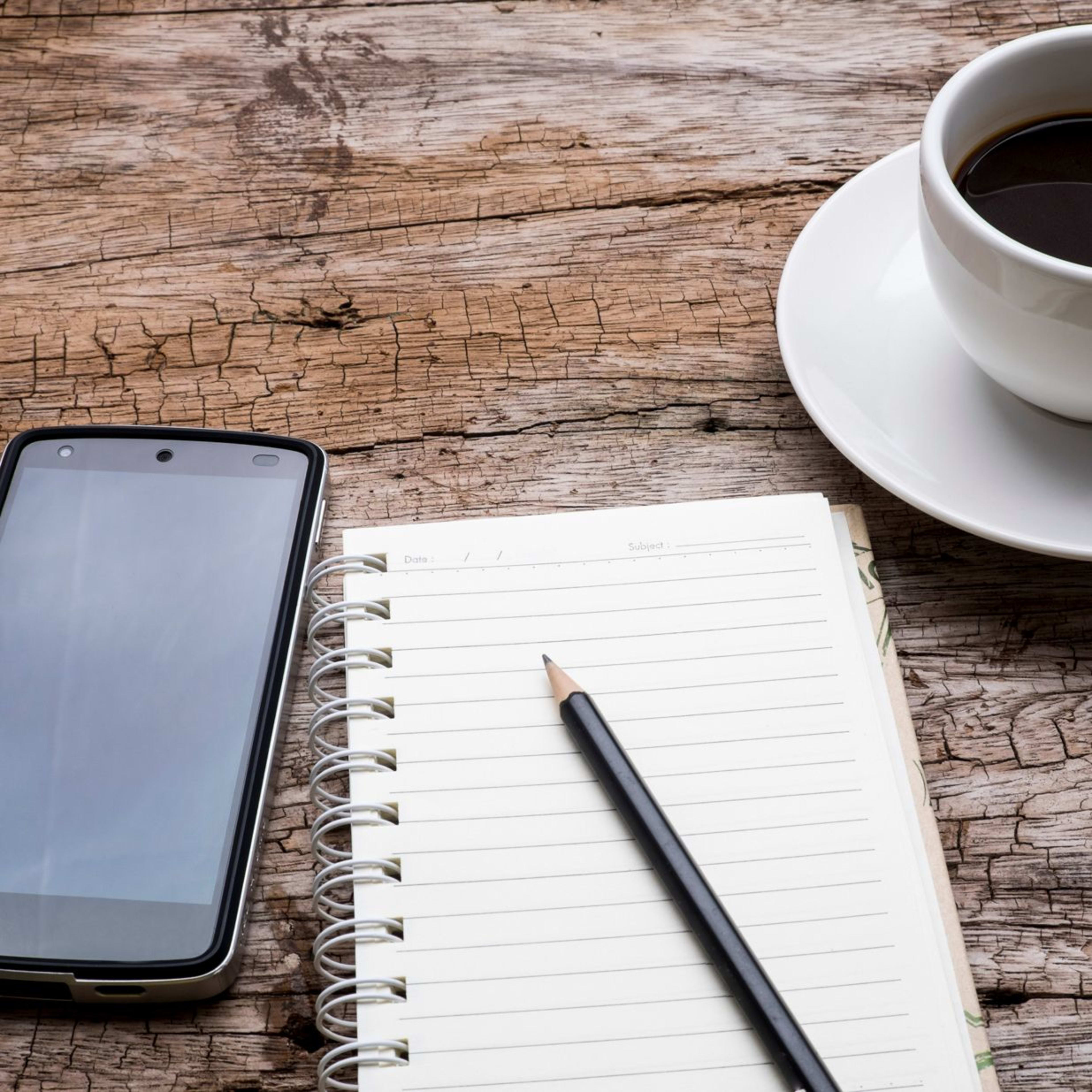 Holztisch mit einem Notizbuch, Smartphone und einer Kaffeetasse