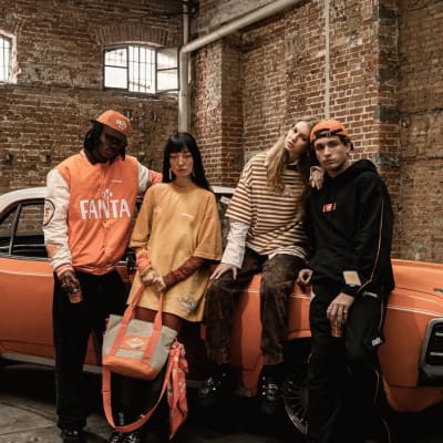 Quatro jovens posam junto a um carro laranja em galpão de tijolos, usando roupas casuais e bonés